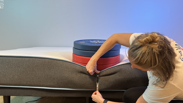 A woman kneeling next to the Bear Original mattress. A stack of weights is perched on the edge, and she's measuring the sinkage with a tape measure.