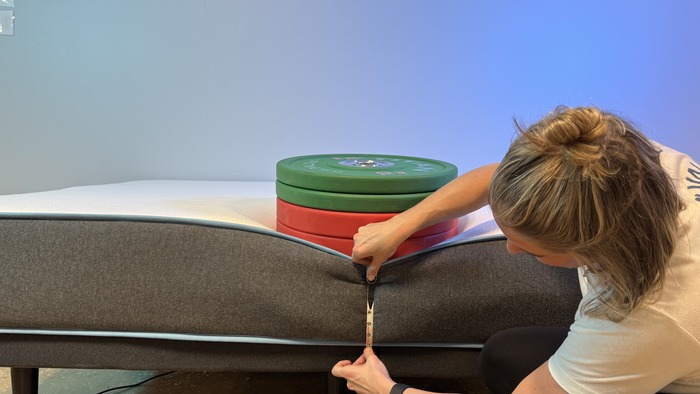 A woman kneeling next to the Bear Original mattress. A stack of weights is perched on the edge, and she's measuring the sinkage with a tape measure.