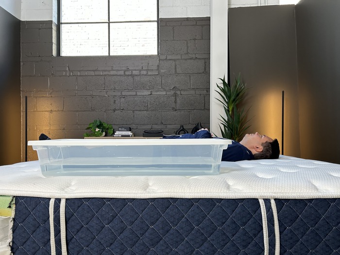 A man lying next to a large bin of water on the Brooklyn Bedding Signature Hybrid mattress in order to test its motion isolation.