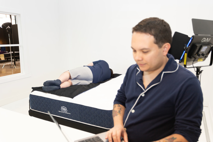 A man holding a laptop while another man behind him rests on a pressure mat on top of the Brooklyn Bedding Signature Hybrid mattress. The man with the laptop is getting a read on the mattress's pressure buildup on the tester's body.