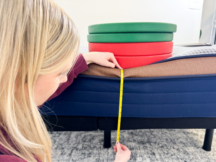 A mattress with a stack of weights on it.  A woman is kneeling next to it with a tape measure to test its edge support.