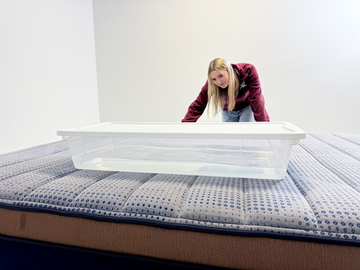 A woman about to get onto a mattress next to a bin filled with water. She'll be able to gauge the mattress's motion-isolating abilities by seeing how much the water moves with her.