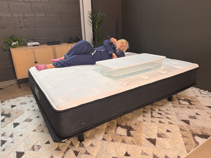A woman lying next to a bin full of water on the Plank Firm mattress. She's rolling around to see how much the water moves, which gives her an idea of well the bed isolates motion.