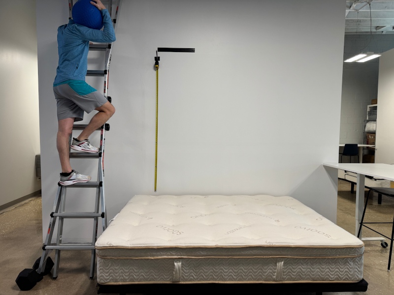 A person climbs a ladder to drop a medicine ball on a Saatva Latex Hybrid.