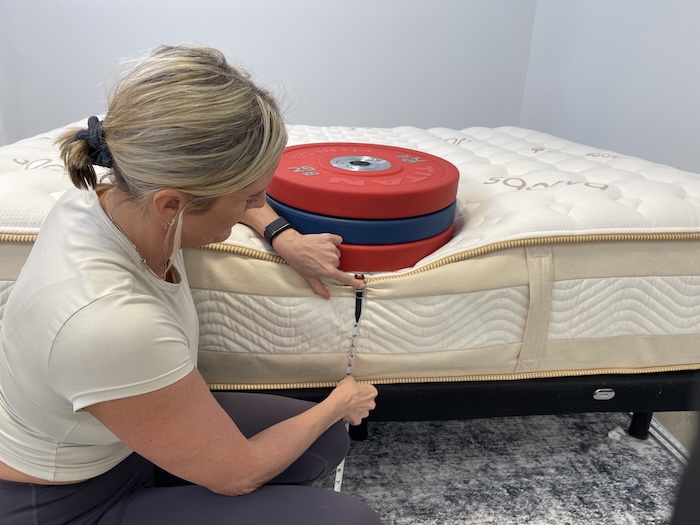 A stack of weights rests on the Saatva Rx. A woman kneels next to them, measuring the mattress's sinkage with a tape measure.