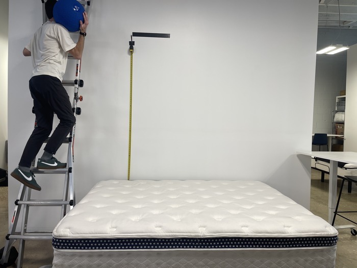A man climbing a ladder with a large blue ball. He's about to drop it on the WinkBed to test the mattress's bounce.