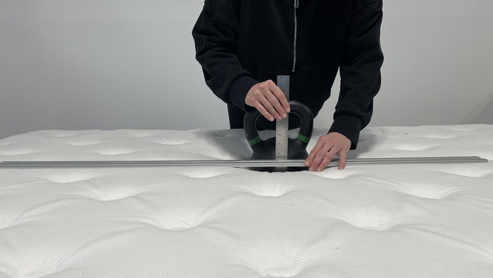 A kettlebell resting on the surface of the WinkBed. Someone is standing next to it and measuring the sinkage with a ruler.
